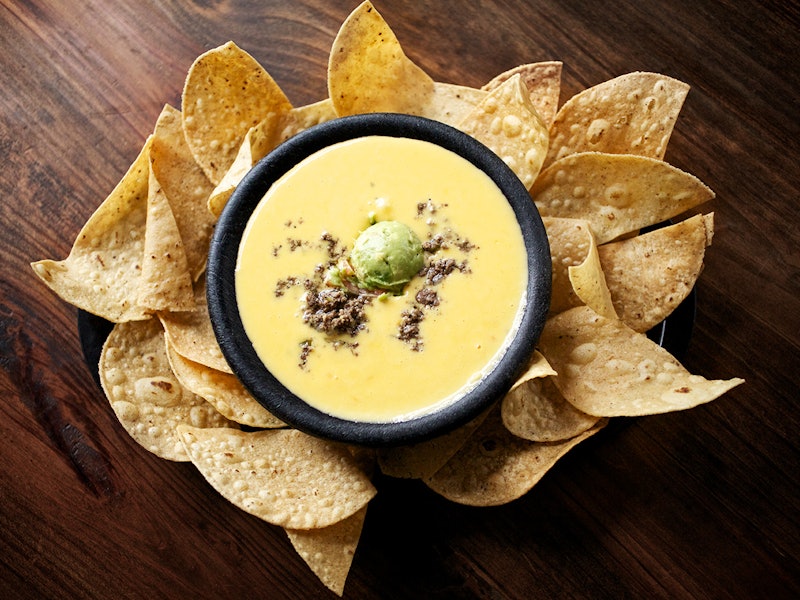 Bowl of yellow melted cheese with a scoop of guacamole, surrounded by corn chips.